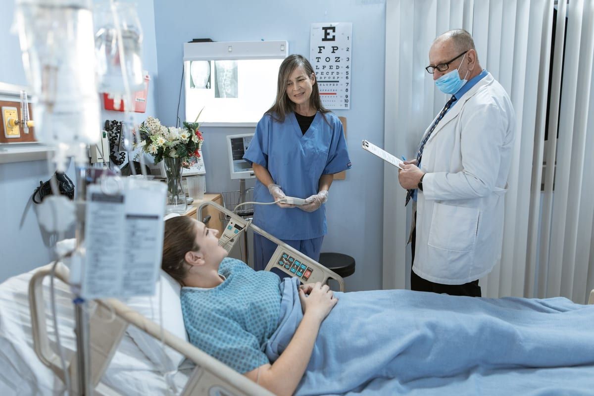 A clinician and a nurse at the bedside of a patient sitting up in a hospital bed, a monitor on the wall behind