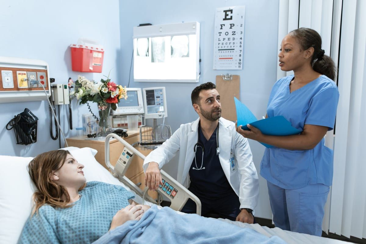 A clinician in a white coat crouching beside a patient in a hospital bed while a nurse in blue scrubs stands over an open blue chart, a cardiac monitor and an eye chart behind them
