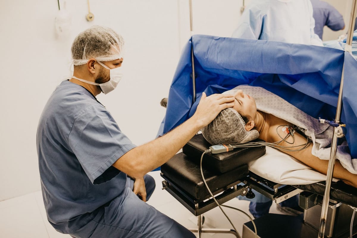 A clinician in blue scrubs, cap and mask crouches at the head of an operating table with a hand on the patient’s forehead, below the blue surgical drape