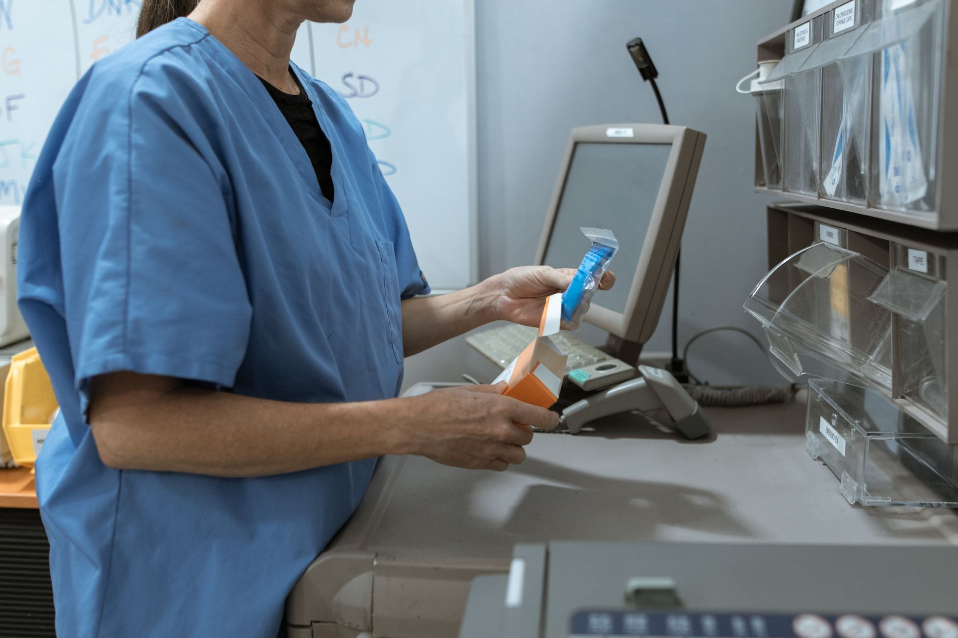 A nurse in blue scrubs at a hospital medication station, opening a boxed medication beside labeled supply bins