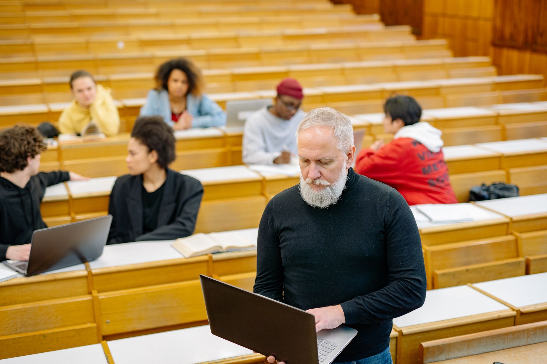 A lecturer holding an open laptop standing among students seated at the tiered wooden desks of a lecture hall