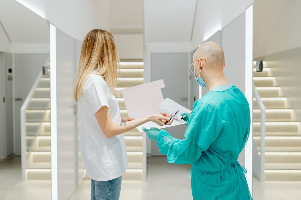 Two clinicians standing in a bright hospital corridor reviewing paperwork on a clipboard together