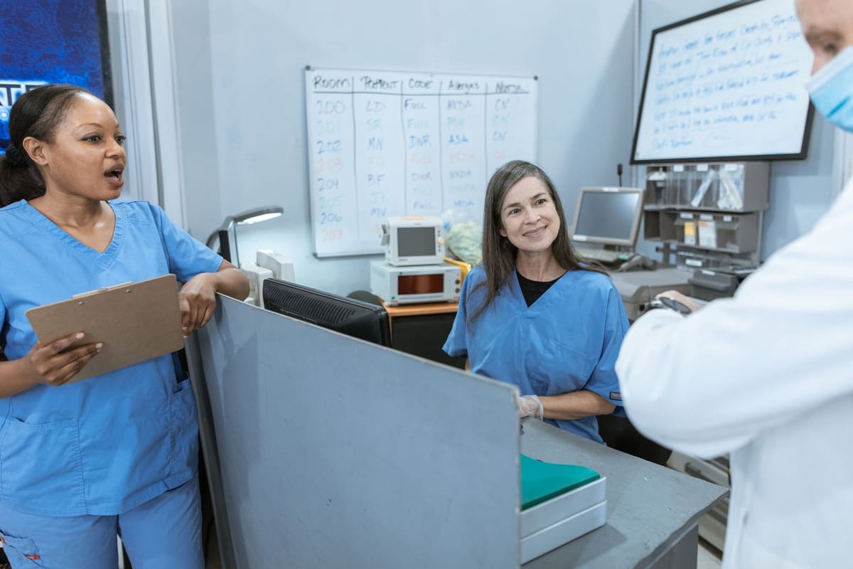 Two nurses in blue scrubs talking across a ward station, a patient assignment board on the wall behind them