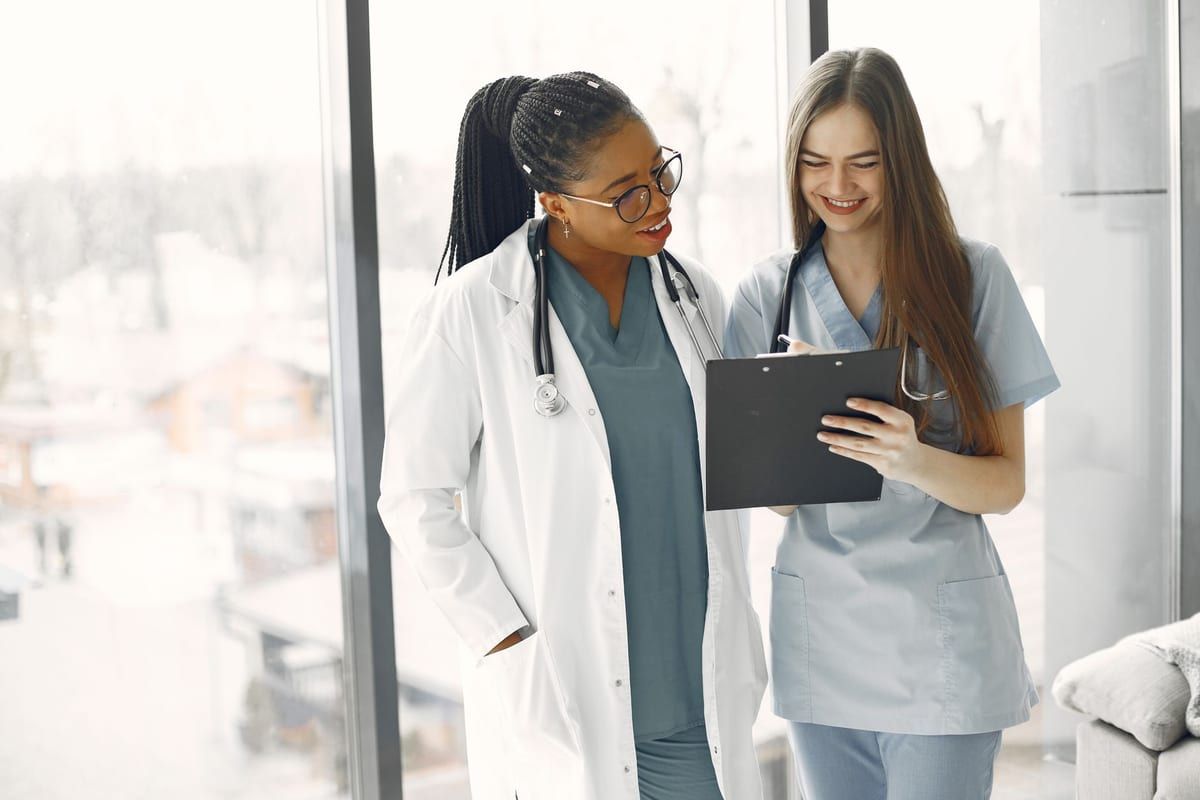 Two clinicians standing at a bright window, one in a white coat over teal scrubs, reading a clipboard together