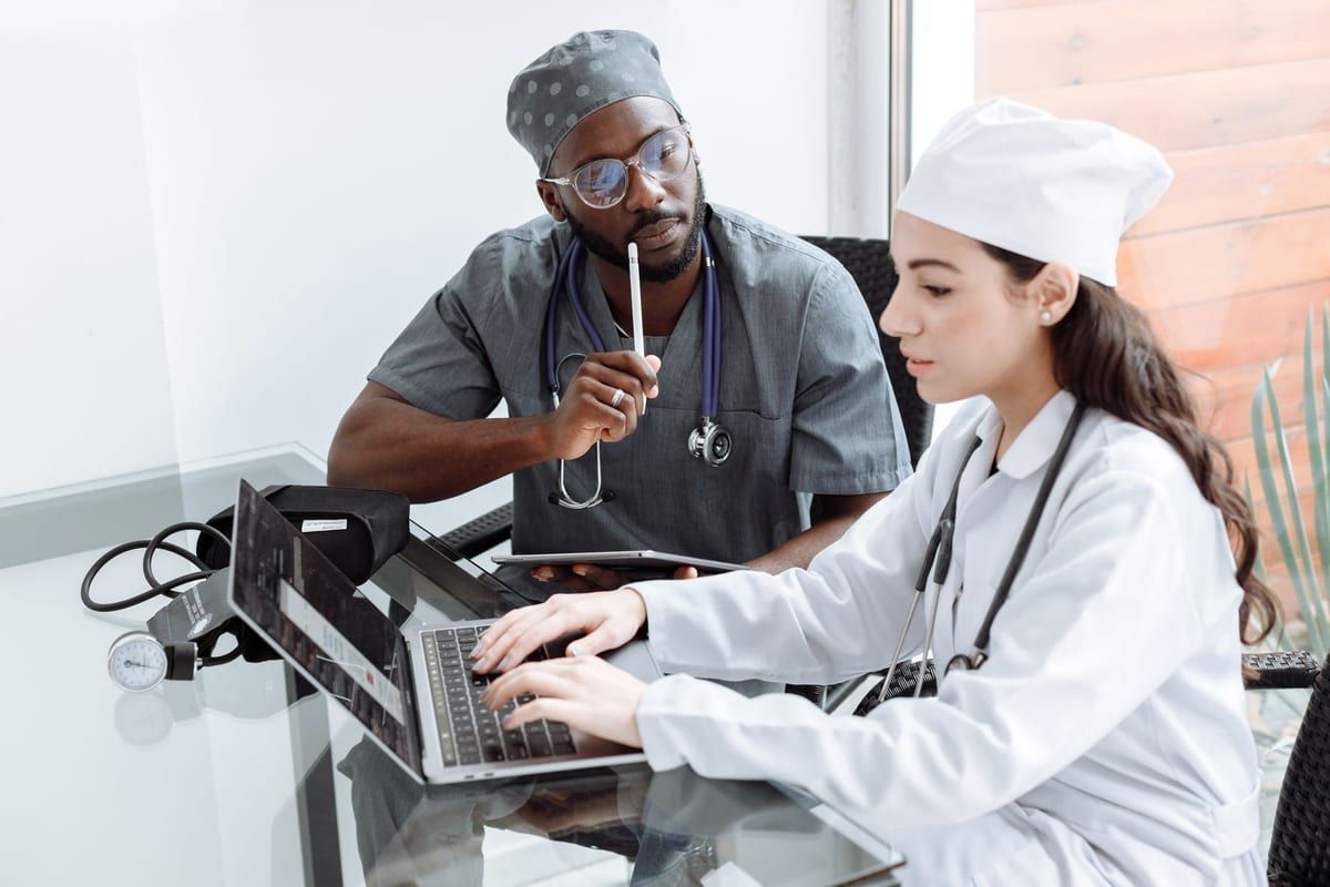 Two clinicians seated at a glass table with a laptop, a tablet and a blood pressure cuff between them, one in scrubs and one in a white coat