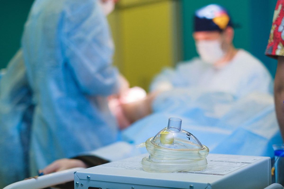 A transparent anaesthesia mask resting on a machine in the foreground, a draped surgical team out of focus behind it