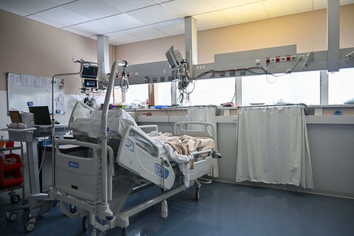 An empty intensive care bay: a hospital bed with its rails up, two patient monitors on an equipment boom, an infusion stand and a curtain along the window