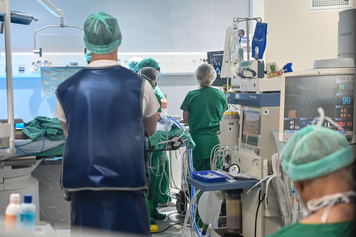 An operating room seen from behind the anesthesia team, a clinician in green scrubs facing an anesthesia machine and a patient monitor while others work at the table