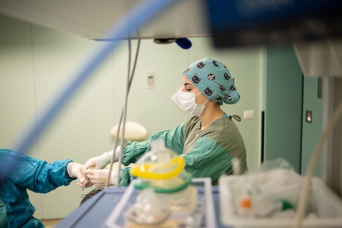 A clinician in a cat-print scrub cap, mask and green gown working at an operating table, with an anesthesia mask and breathing circuit in the blurred foreground