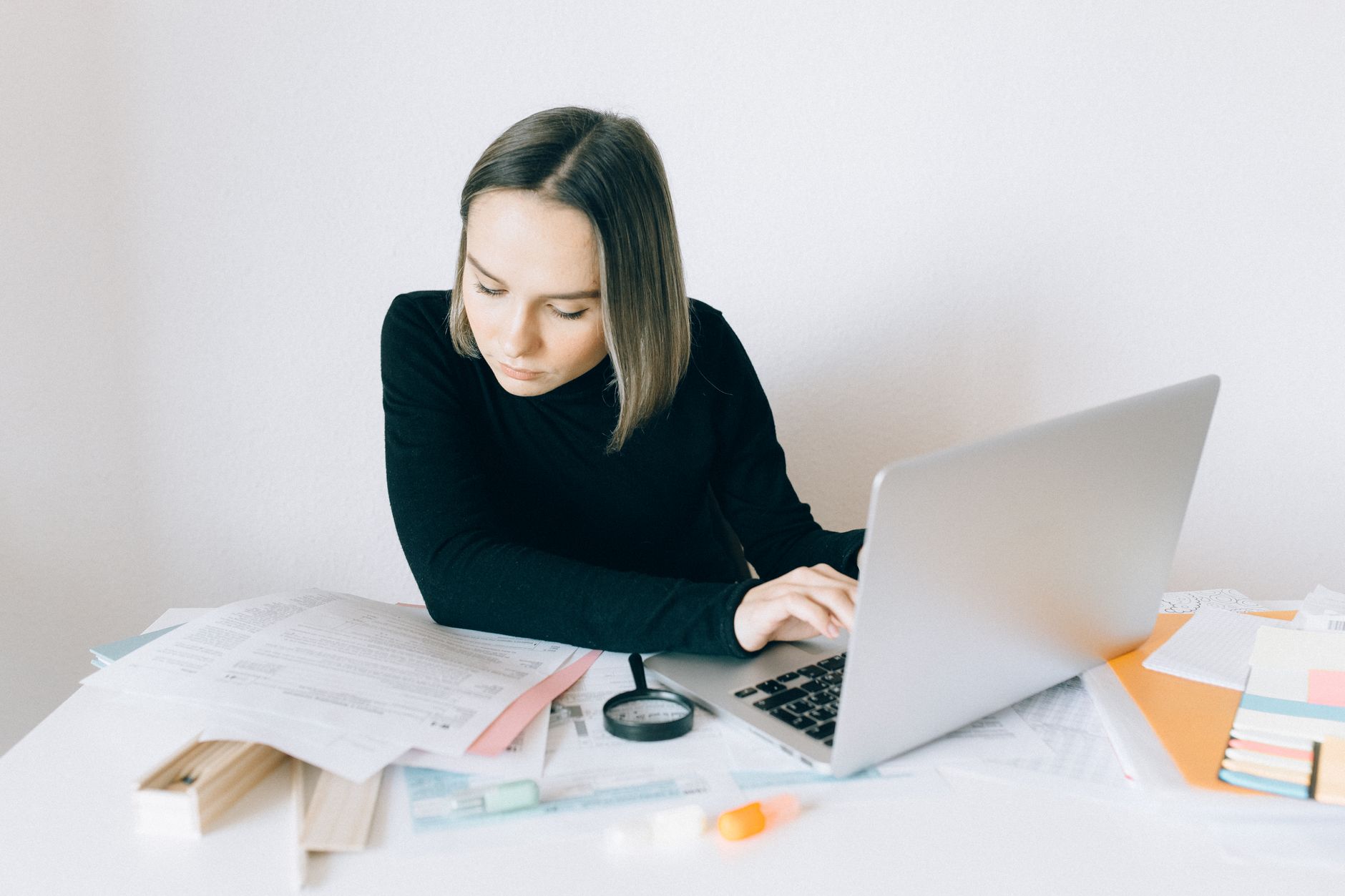 A woman working at a laptop at a white desk covered in printed forms, a magnifier and colored folders