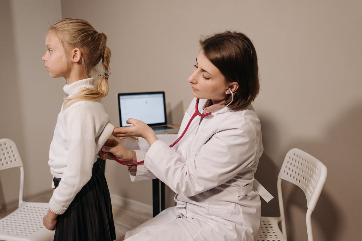 A clinician with a red stethoscope listening to the back of a school-age girl standing beside her, an open laptop on the desk behind