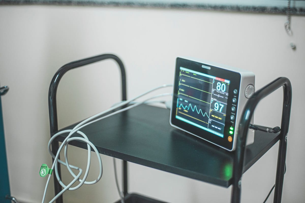 A patient monitor on a black wheeled cart showing a heart-rate waveform and vital-sign readings, its sensor cables looped beside it