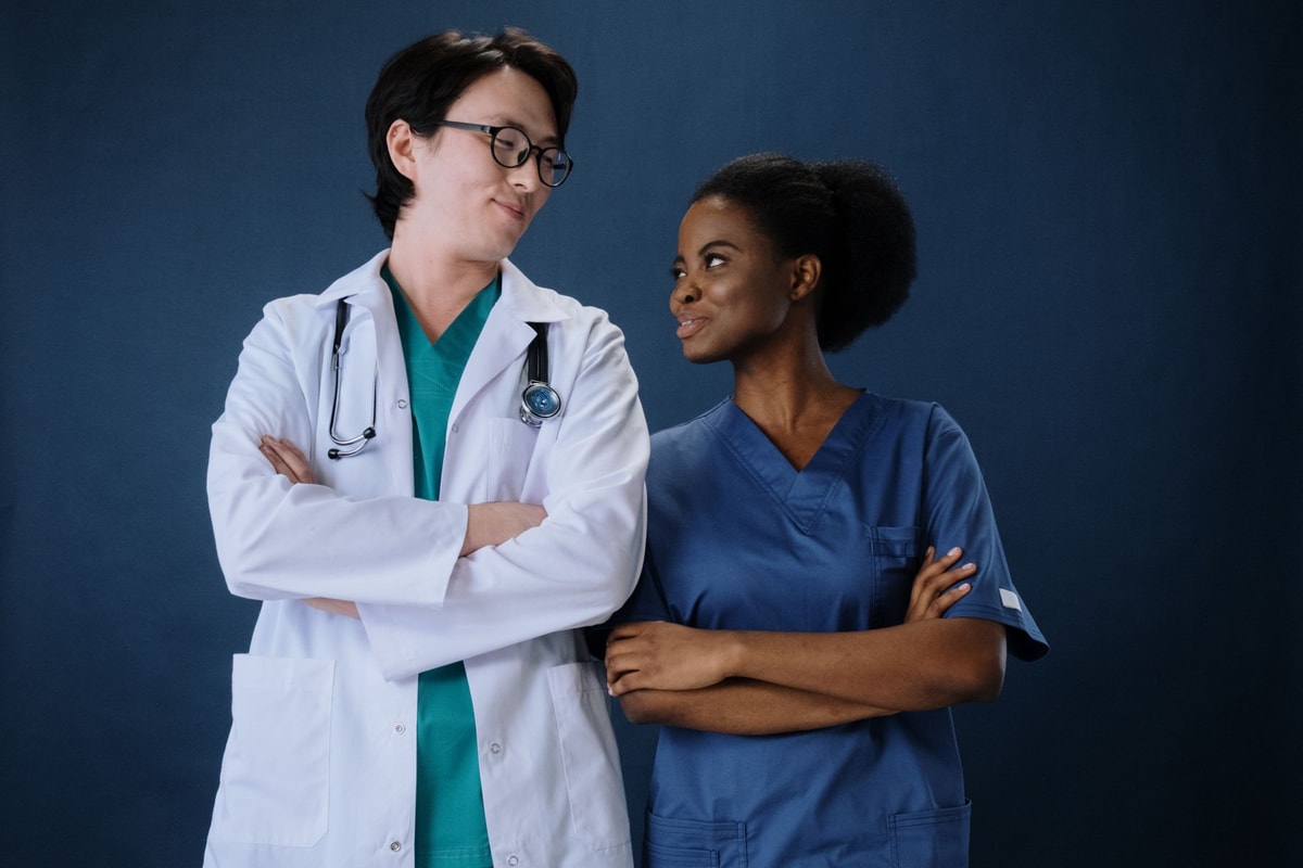 A man in a white coat with a stethoscope over teal scrubs and a woman in blue scrubs standing side by side with their arms folded, smiling at each other against a navy backdrop