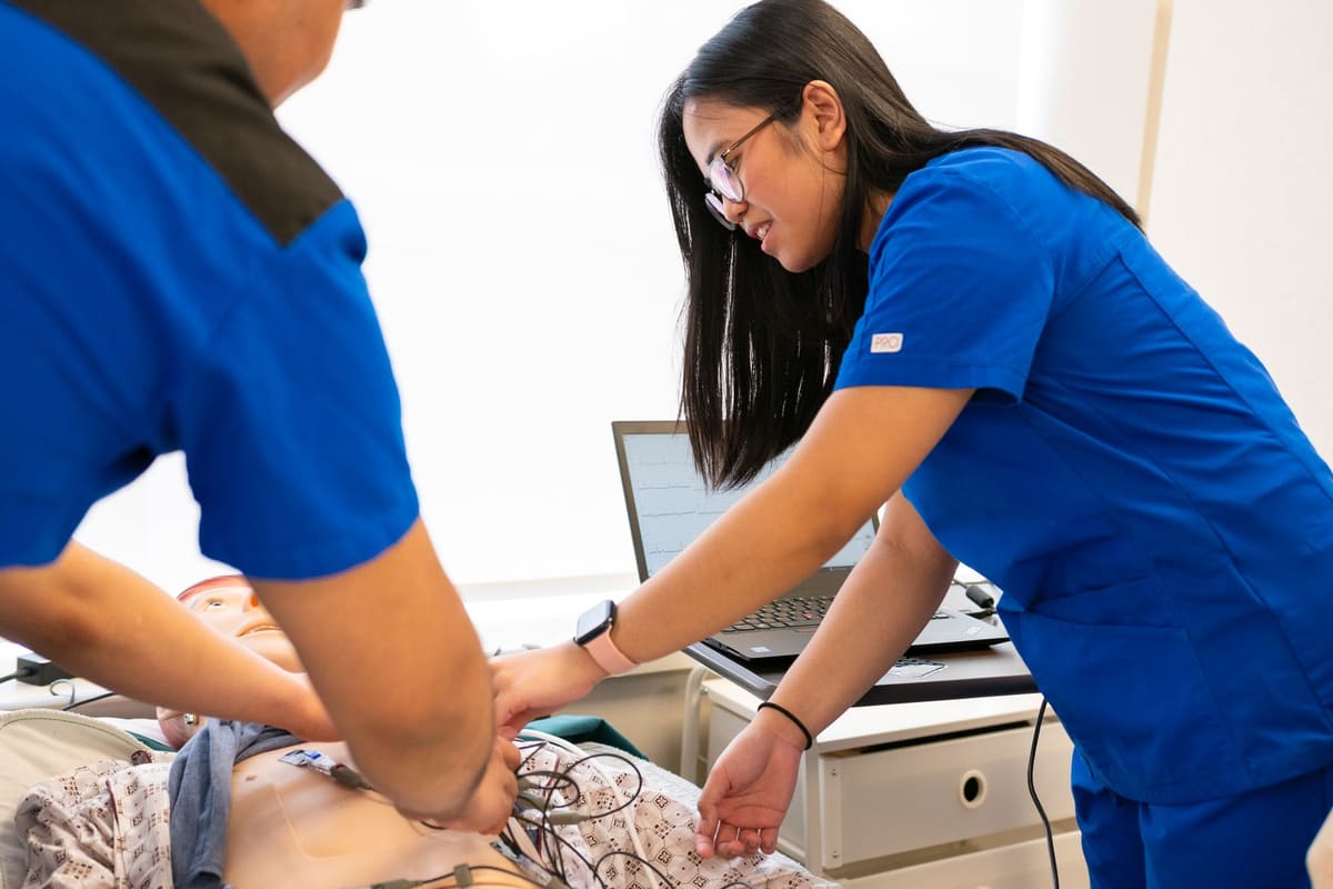 Two nurses in blue scrubs practicing on a training manikin wired to a laptop showing an ECG trace