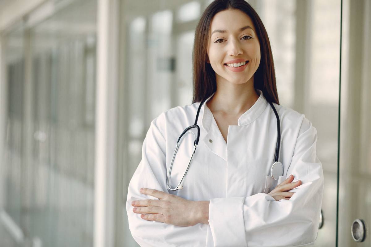 A clinician in a white coat with a stethoscope, arms folded, in a bright clinic corridor