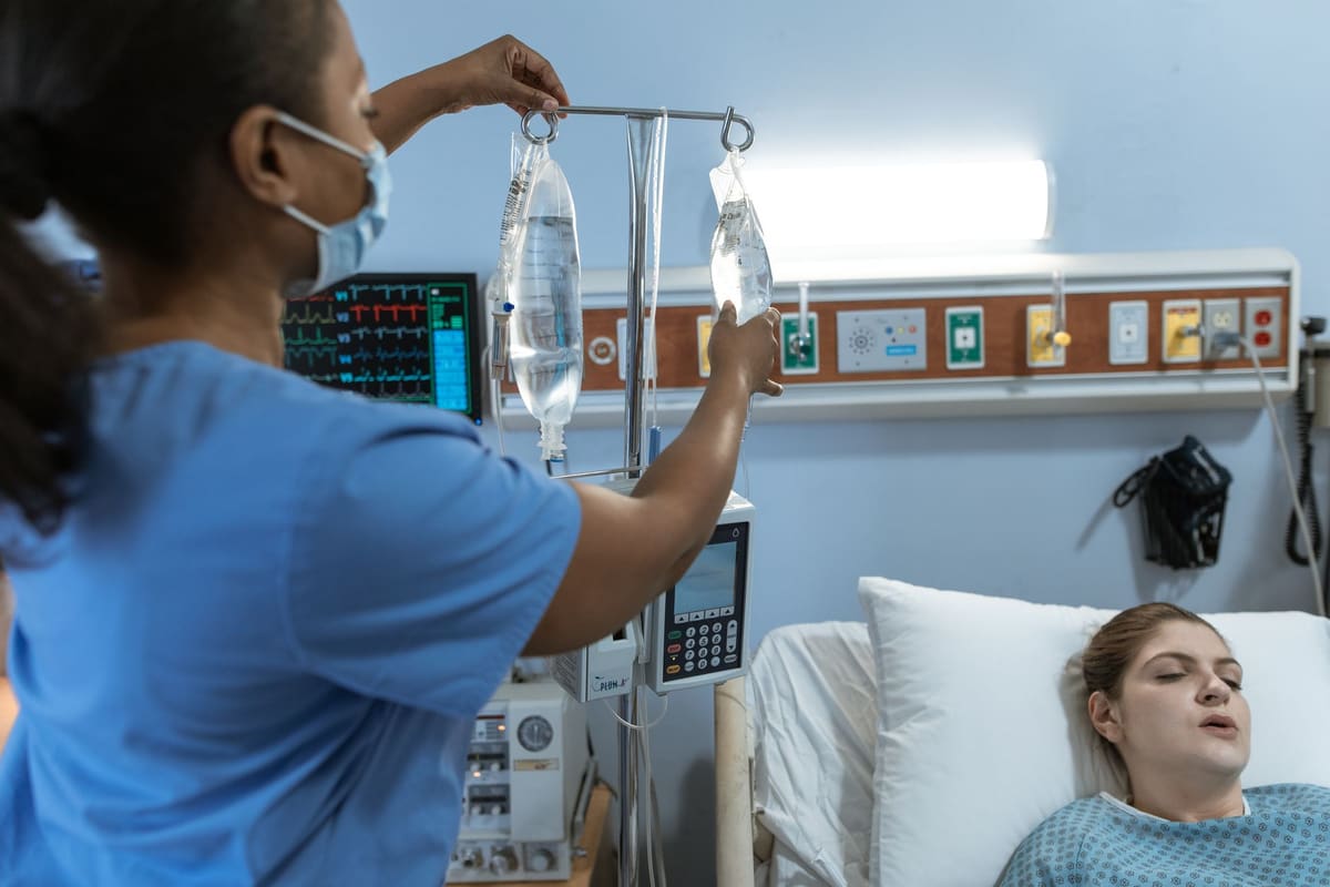A nurse in blue scrubs hanging a bag of intravenous fluid on a stand beside a patient in a hospital bed, a cardiac monitor behind