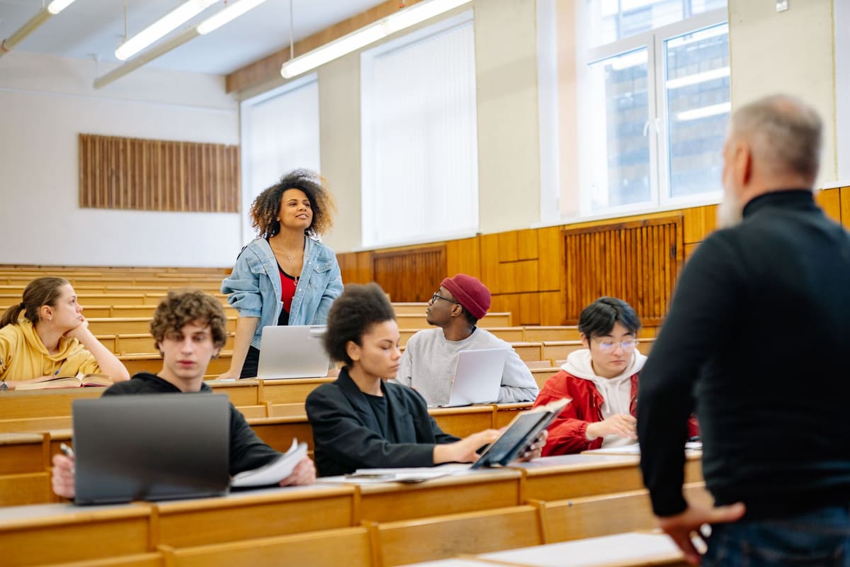 A lecturer standing at the front of a tiered lecture hall as students at wooden desks look up from laptops and notebooks