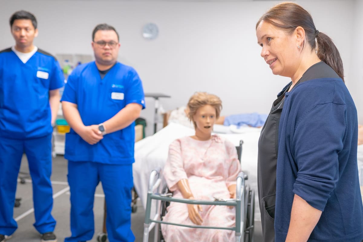 An instructor talking with two students in blue scrubs in a simulation lab, a training manikin seated in a wheelchair behind them