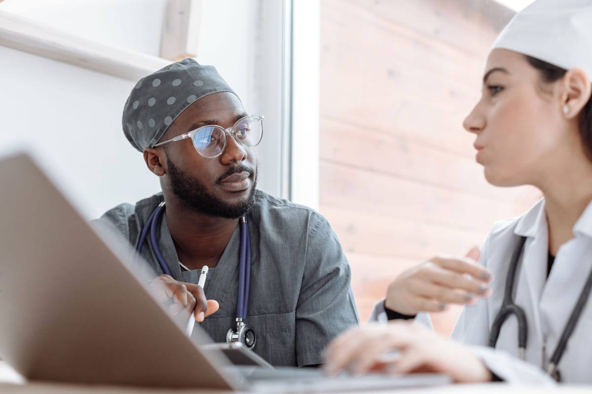 A clinician in gray scrubs and a surgical cap, a stethoscope around his neck, listening to a colleague in a white coat as they work at a laptop by a window