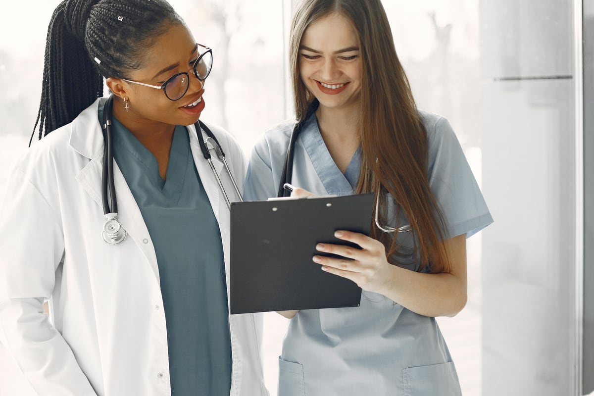 A clinician in a white coat over teal scrubs and a nurse in pale blue scrubs reading the same clipboard, both smiling, in a window-lit corridor