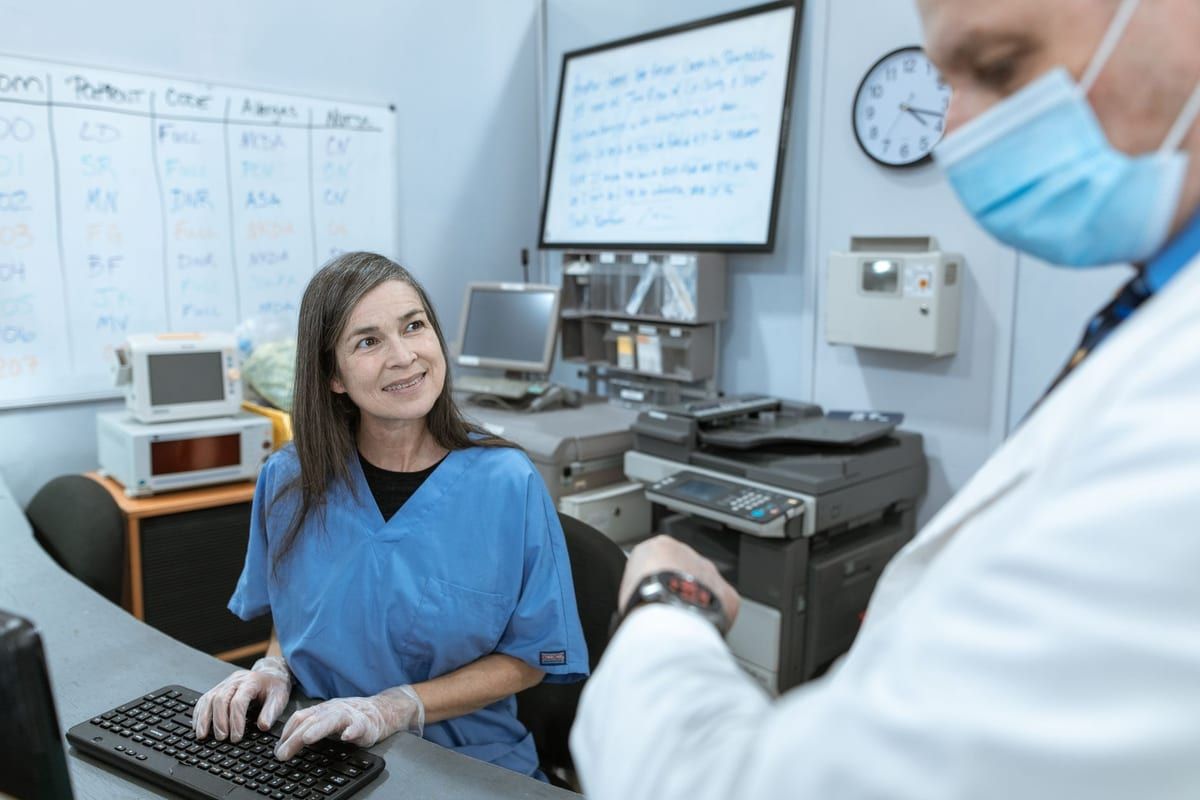 A nurse in blue scrubs at a ward station keyboard looking up at a colleague in a white coat, a room-by-room assignment whiteboard on the wall behind