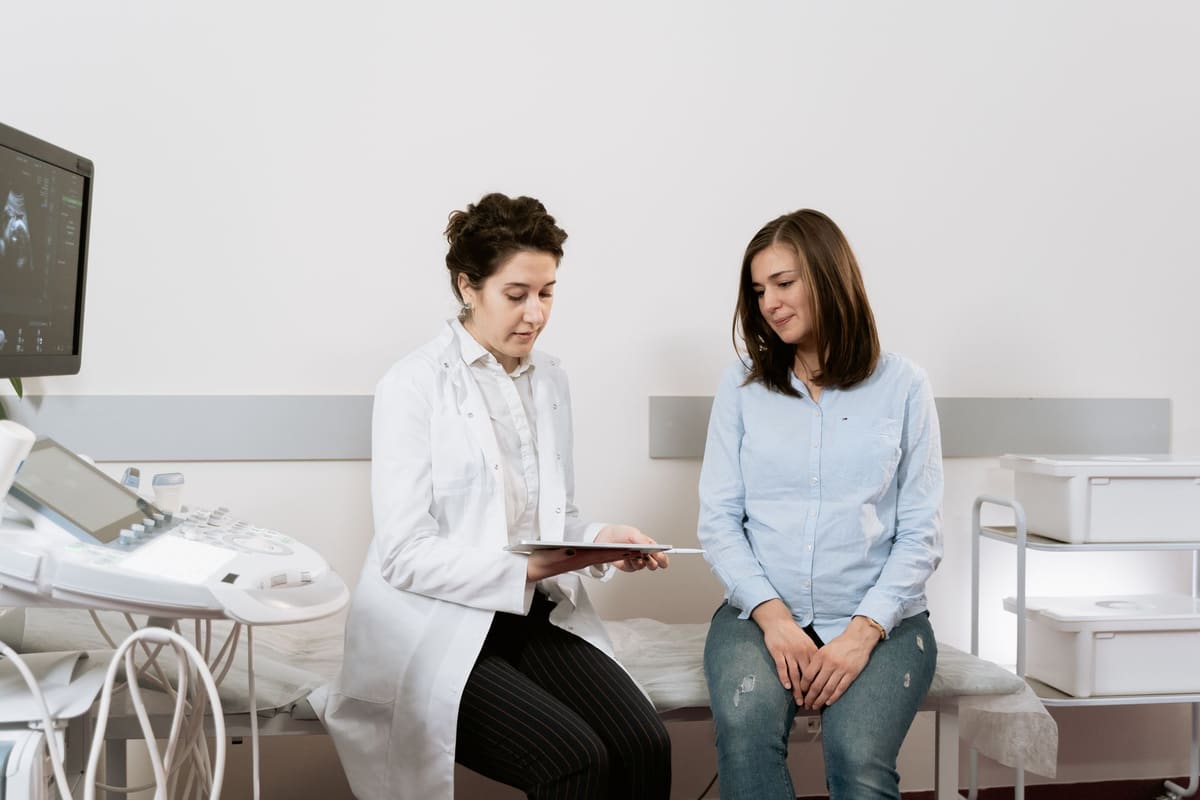 A clinician in a white coat seated beside a patient in an exam room, an ultrasound machine at her side