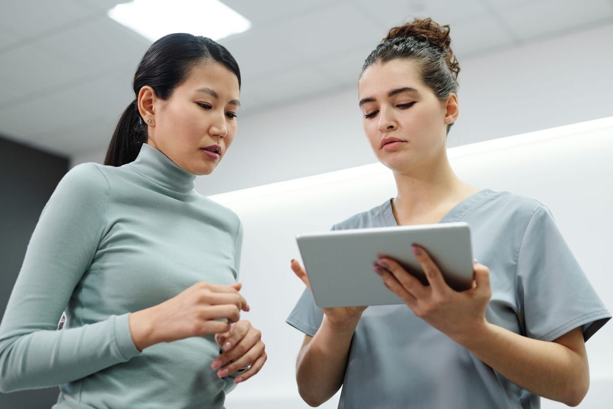A clinician in gray scrubs holding a tablet and showing it to a woman standing beside her in a bright clinic room