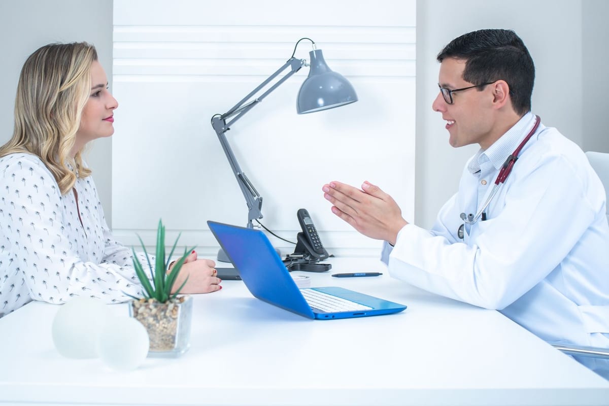 A clinician in a white coat and stethoscope talking across a desk with a seated patient in a bright consulting room, a laptop and a desk lamp between them