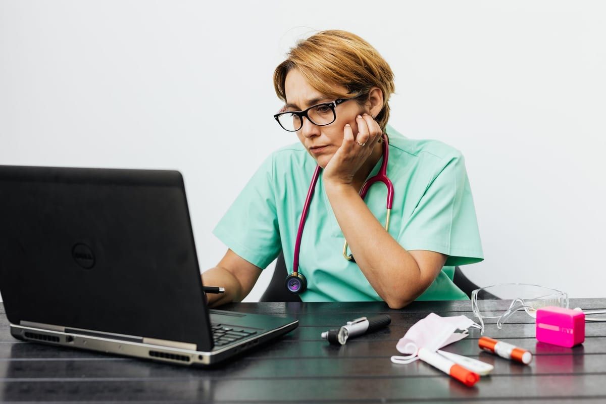 A nurse in mint scrubs and glasses working at a laptop, chin resting on one hand, a stethoscope and instruments on the desk