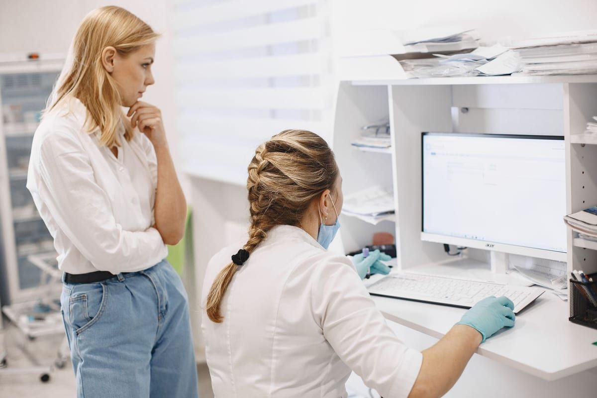 A clinician in a white coat seated at a records terminal at a clinic desk, a colleague standing beside her looking at the screen