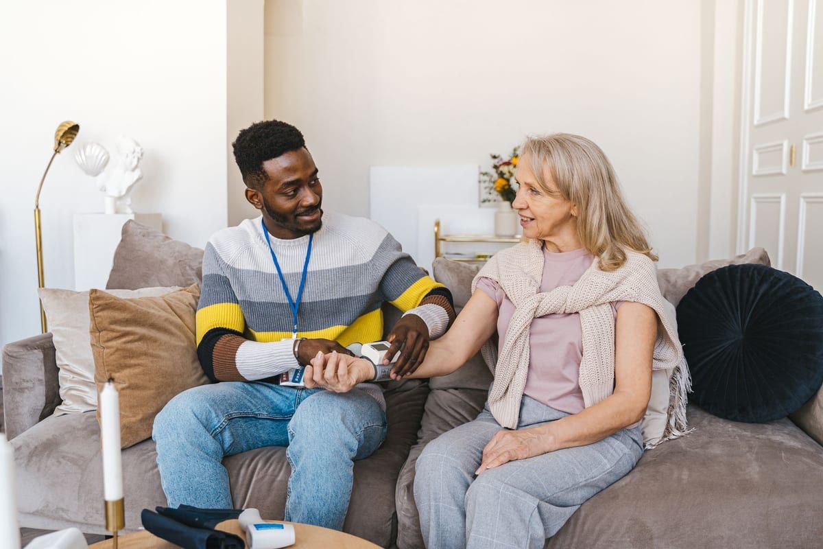 A health worker in a striped jumper and lanyard sitting on a sofa beside an older woman, taking her blood pressure with a wrist monitor