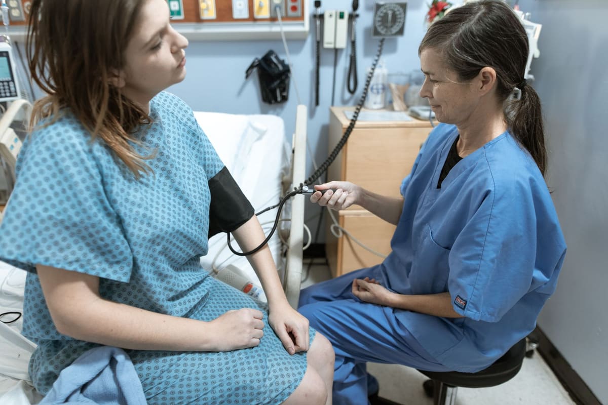 A nurse in blue scrubs seated beside a patient in a gown, taking her blood pressure in an exam room