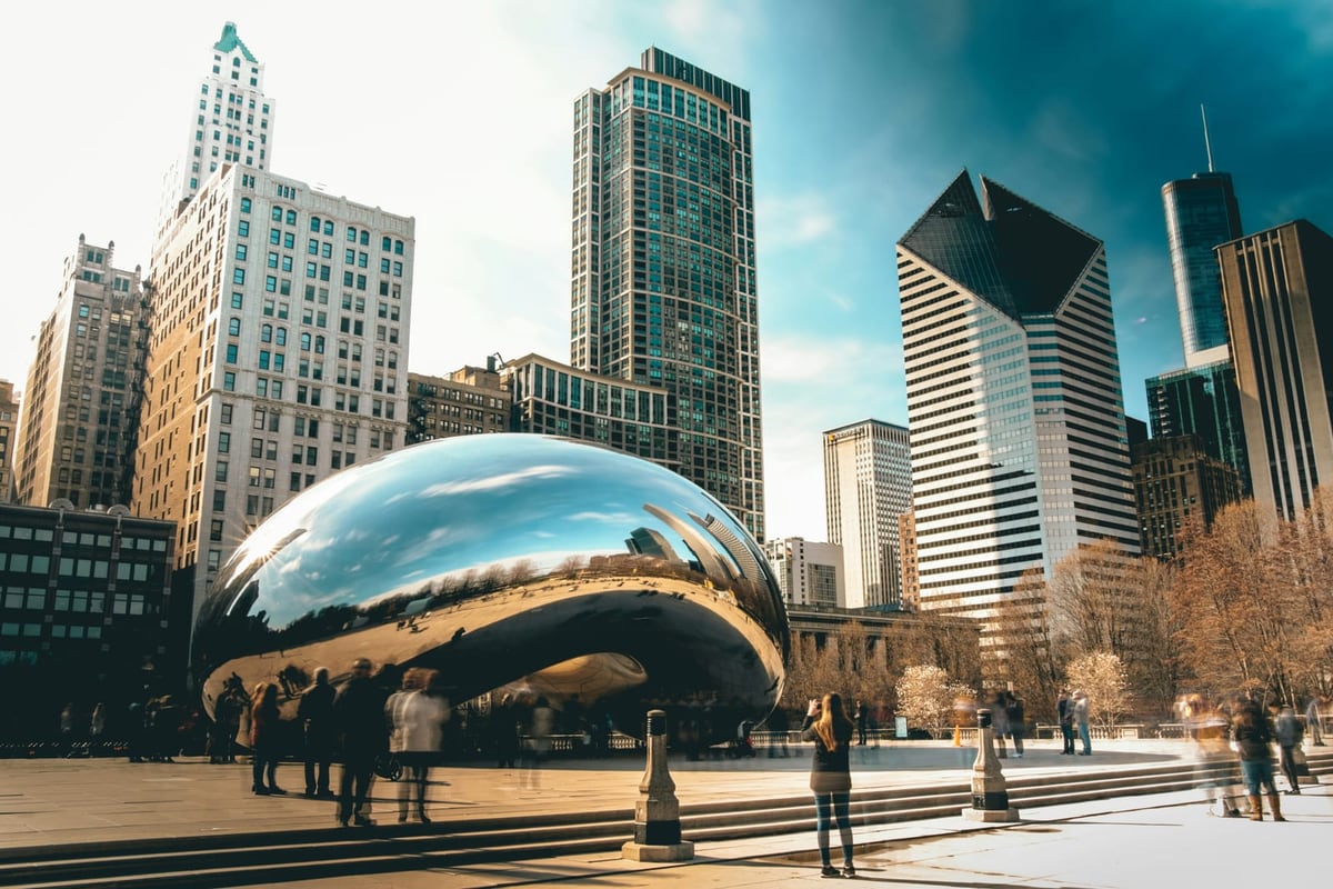 A polished steel sculpture shaped like a bean reflecting a city skyline, with people gathered around it in a plaza