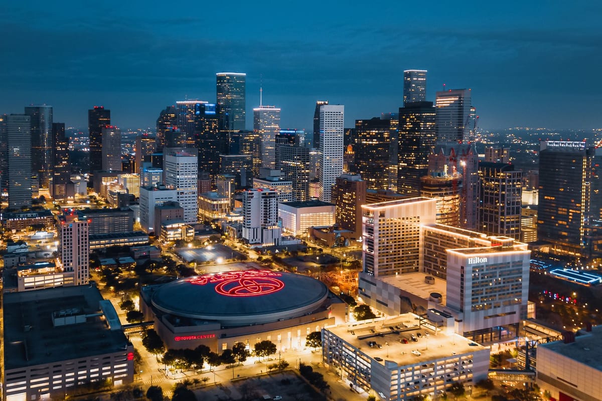 A dense downtown lit at night, seen from above, with an arena roof and lit streets in the foreground