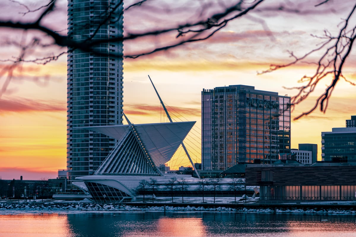 A white winged structure on a lakefront beside glass towers at sunset, framed by bare branches, with ice on the water