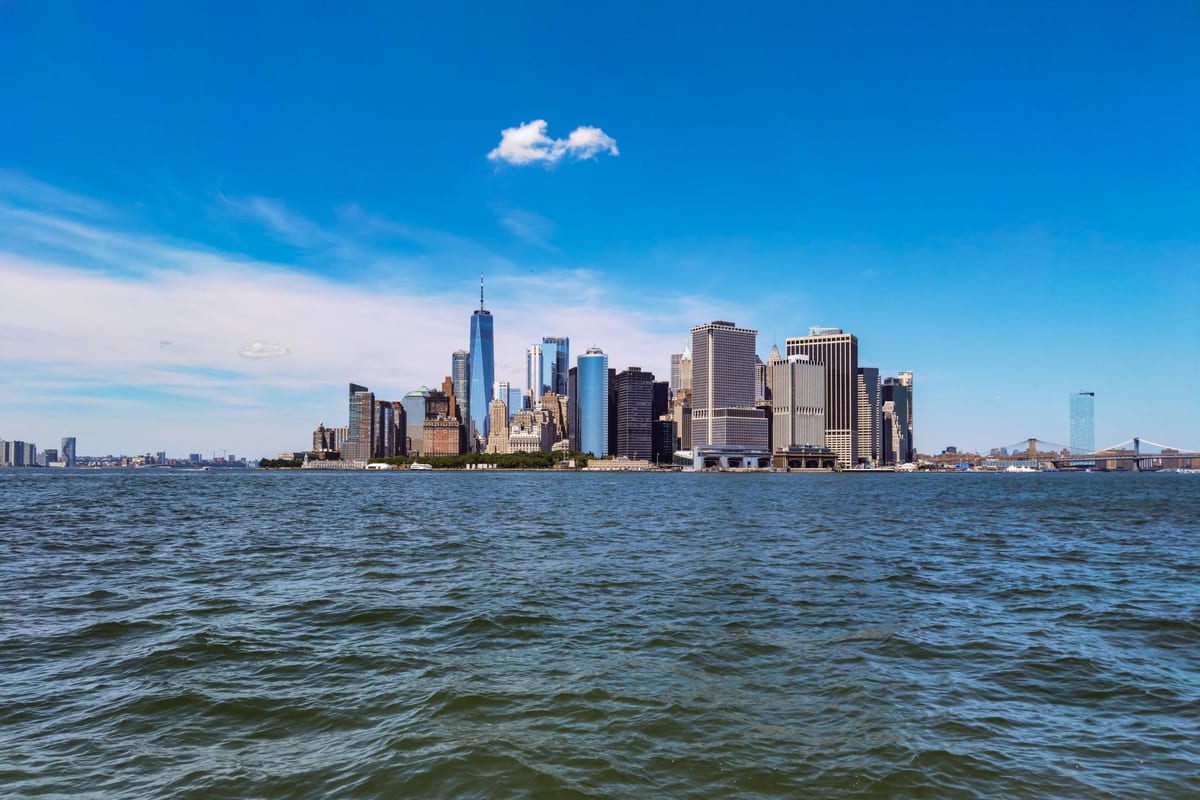 A dense cluster of towers on the tip of an island seen across open water under a deep blue sky, a suspension bridge at the right edge