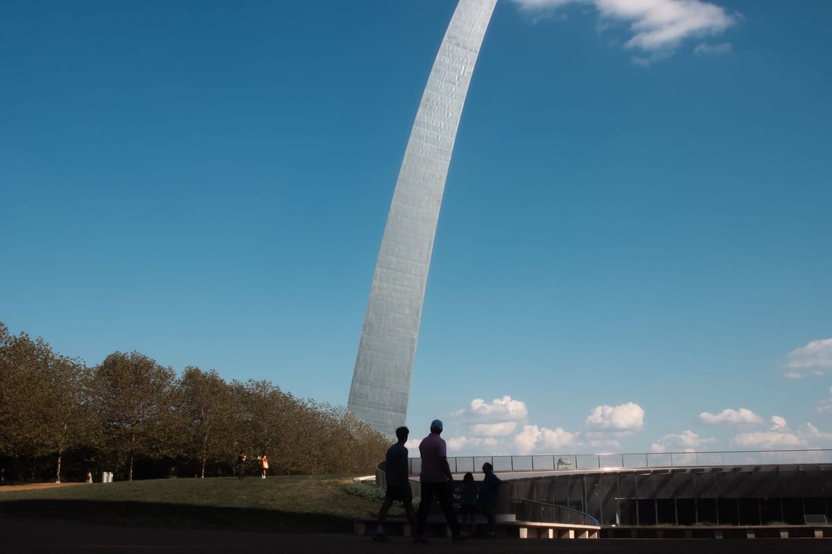 A tall stainless steel arch rising out of parkland against a blue sky, with people walking a path beneath it