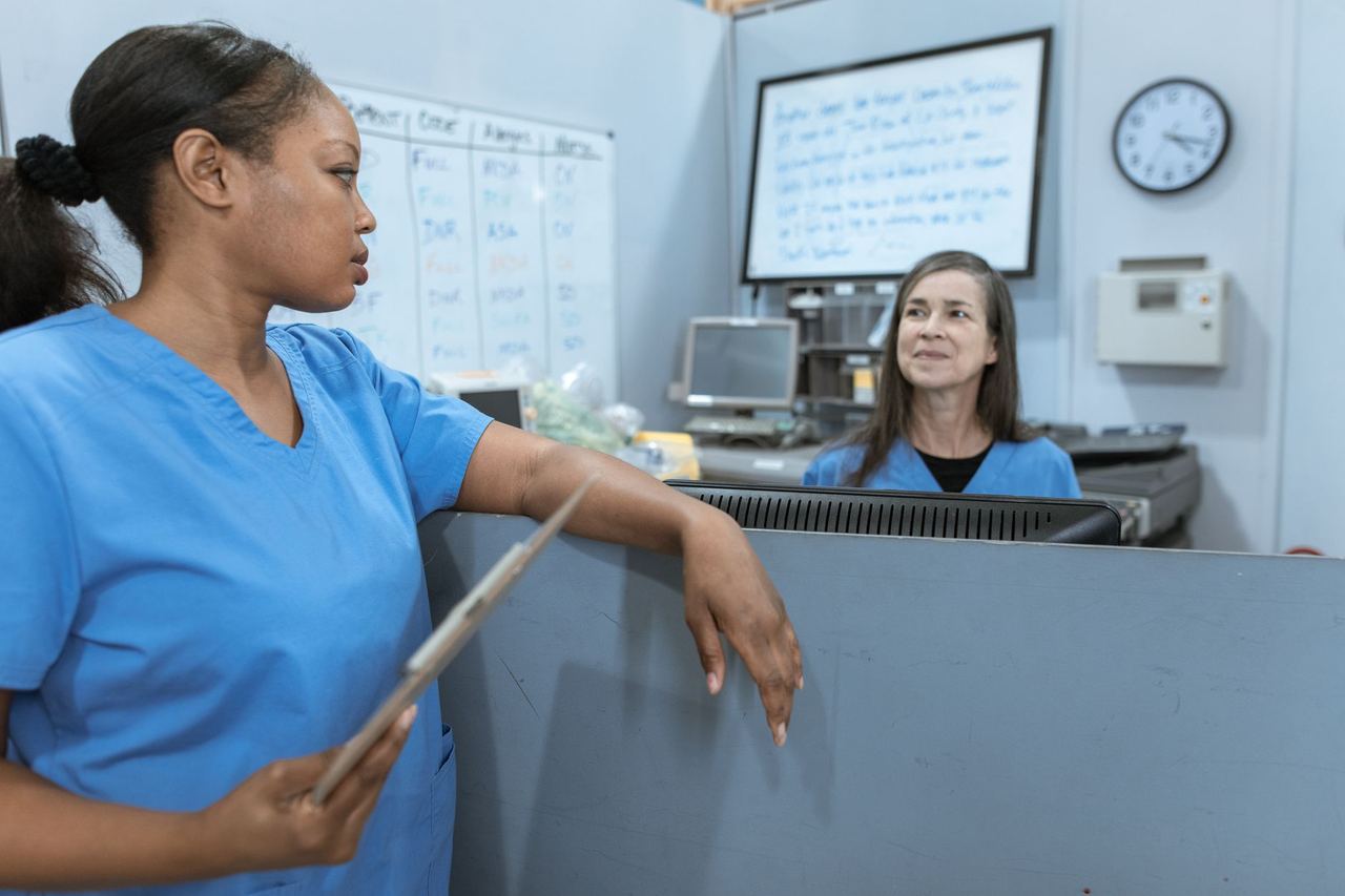 Two nurses in blue scrubs talking across a nurses' station, one holding a clipboard, a shift whiteboard behind them