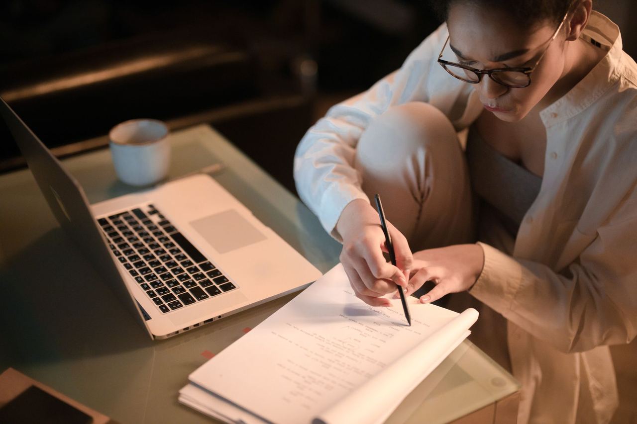 A student working at night at a glass desk, writing notes beside an open laptop and a mug