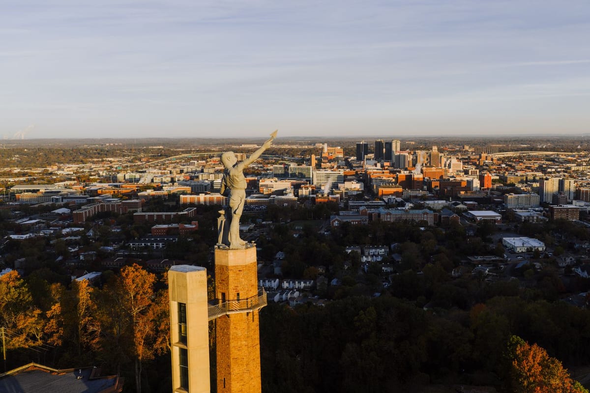 A cast-iron statue on a wooded hilltop above a city skyline at sunset