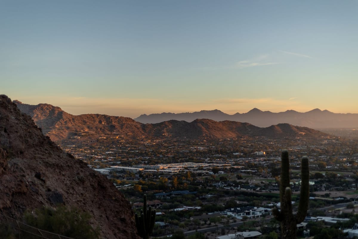 A low desert city spreading to the horizon below dark mountains at sunset, cacti in silhouette