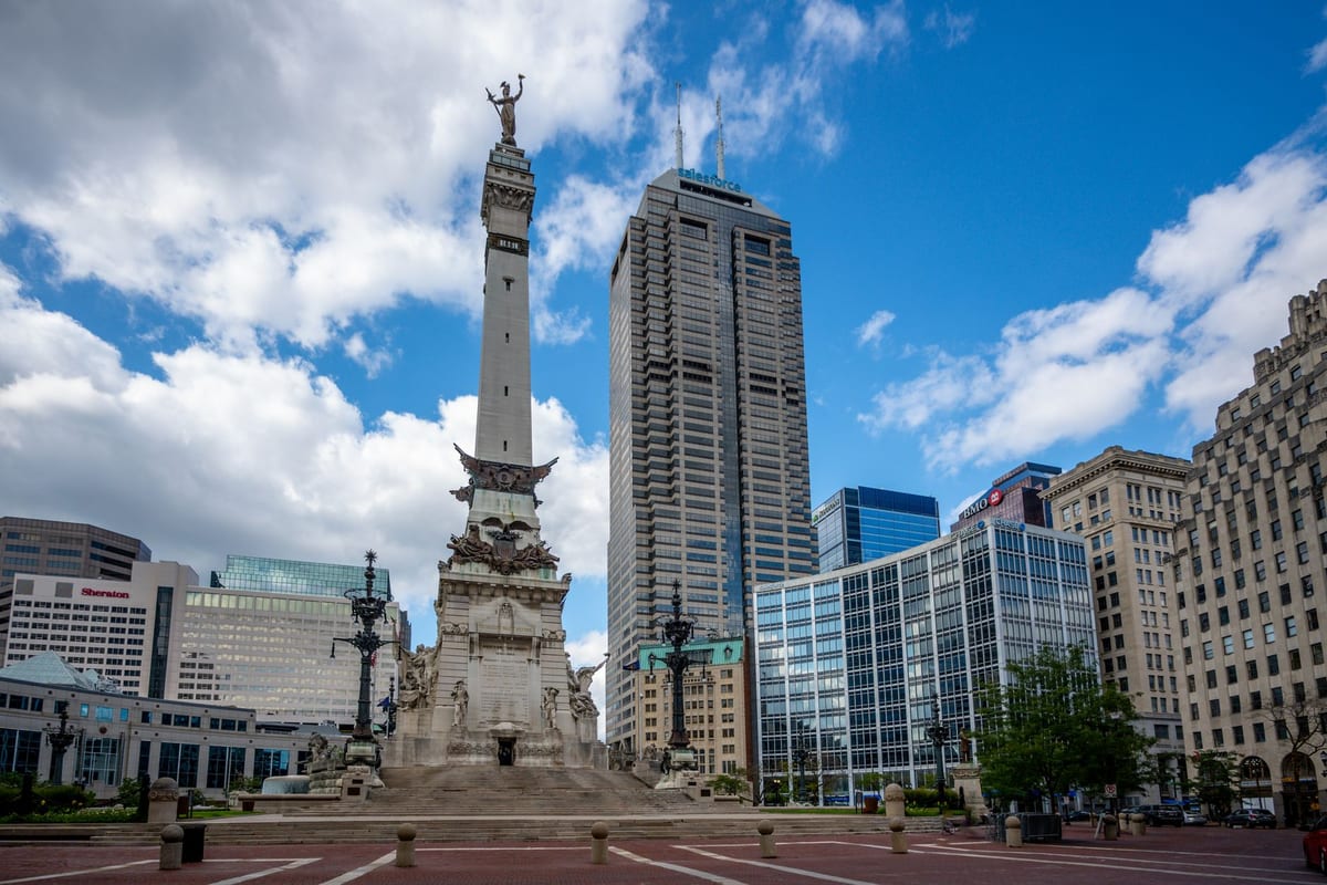 A tall stone monument on a circular plaza with office towers behind it under a blue sky