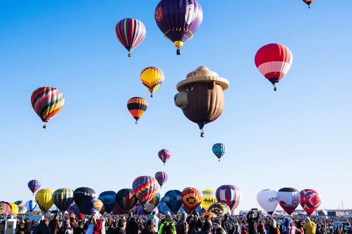 Dozens of hot air balloons in patterned colors rising over a city at dawn