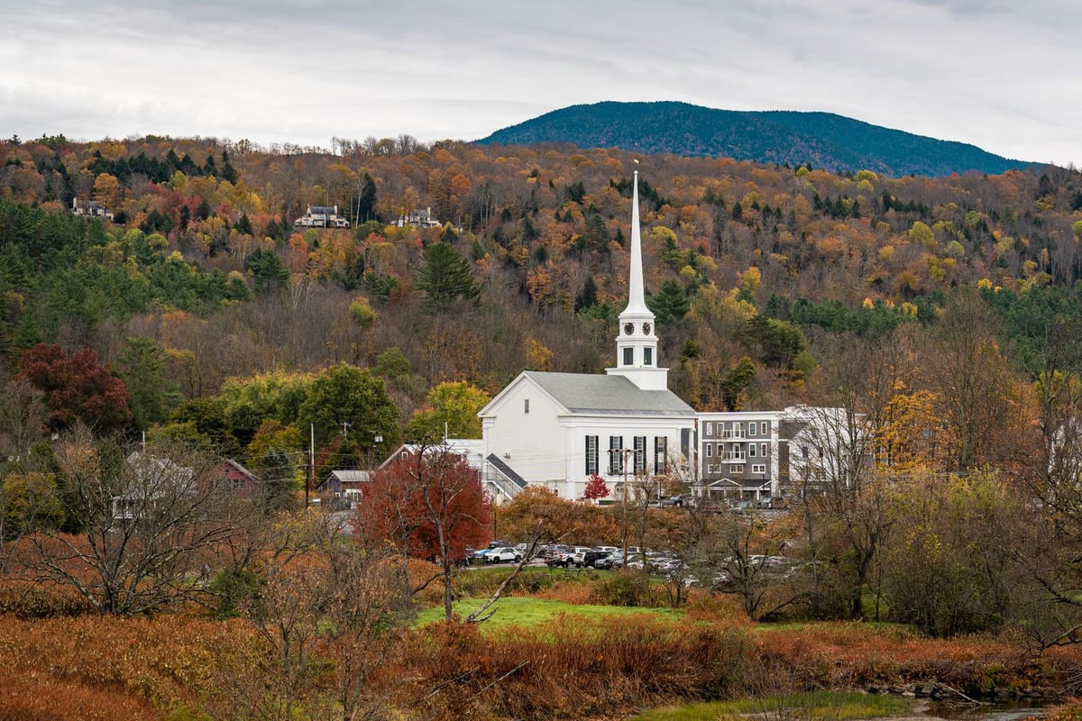 A white clapboard church with a tall steeple among autumn trees below a wooded hill